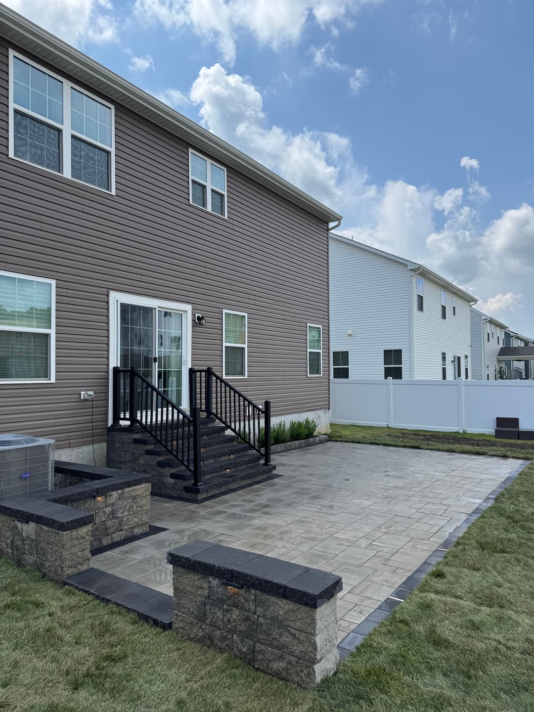 Modern home exterior with stone patio, black steps, and landscaped yard under a cloudy sky.