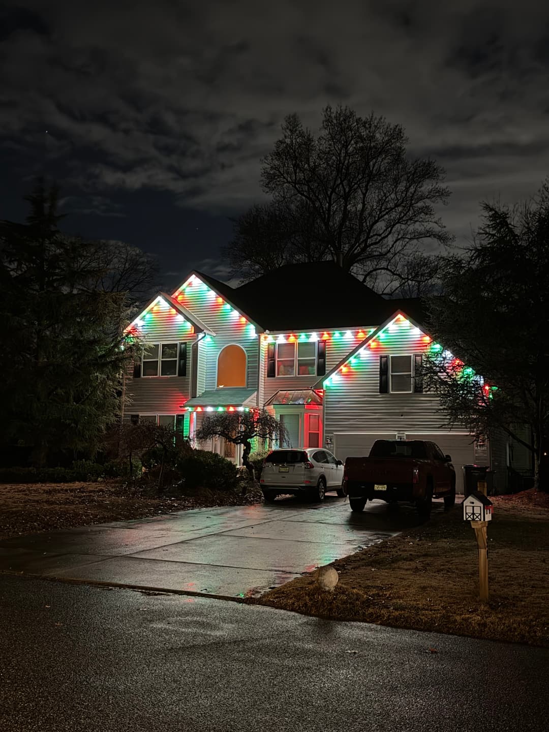 House decorated with colorful Christmas lights at night, surrounded by trees and a cloudy sky.