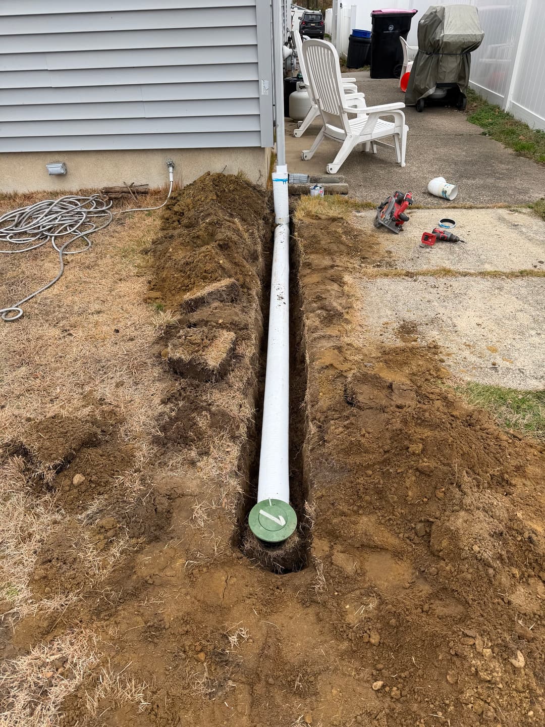 Underground drainage pipe installation in a yard with tools and chairs nearby.