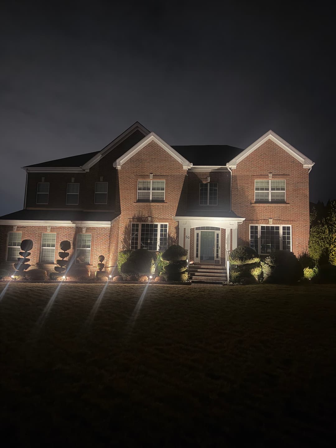 Illuminated brick house at night with landscaped yard and decorative topiary.