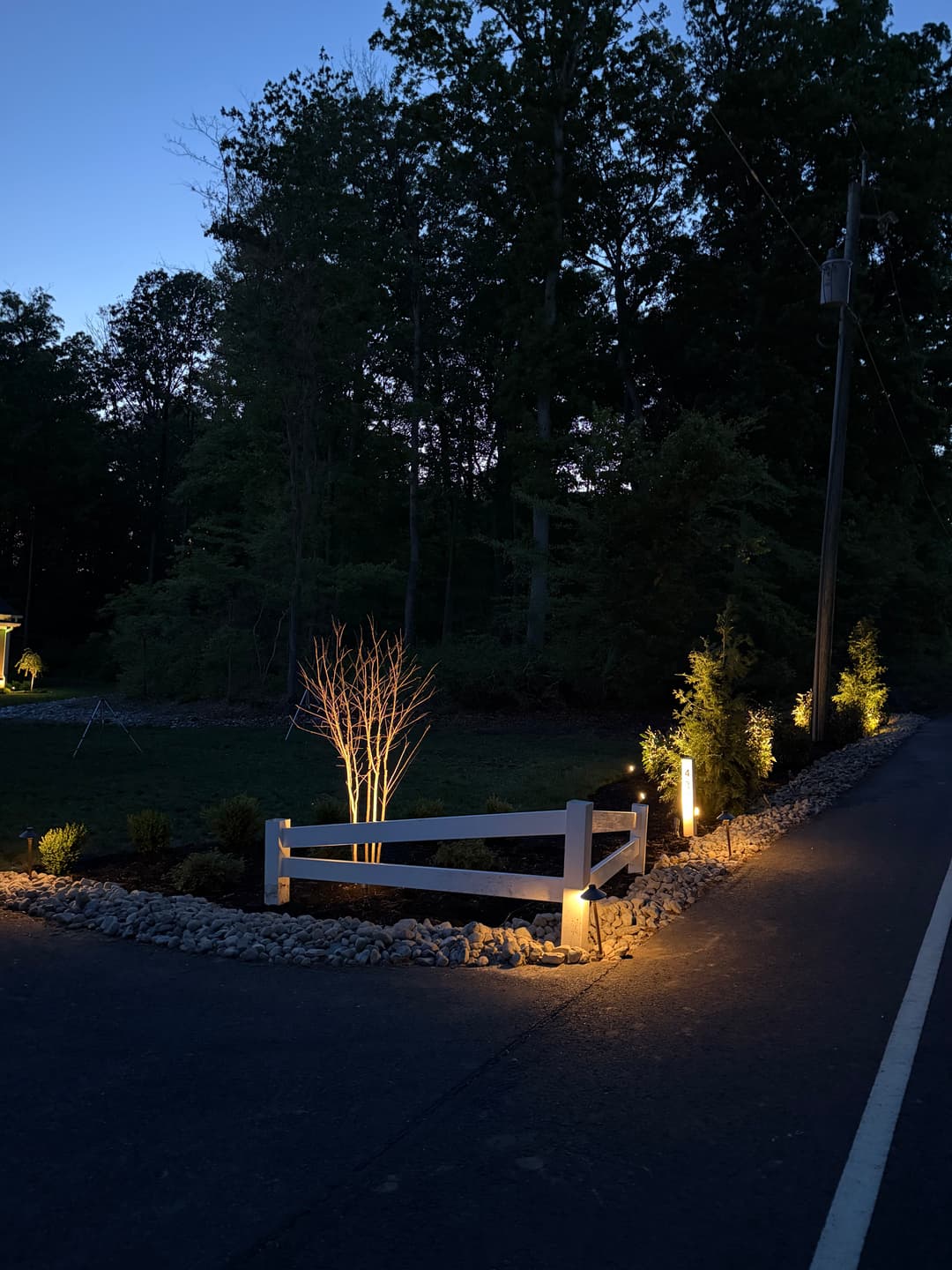 Illuminated pathway bordered by white fence, trees, and landscaping at dusk.