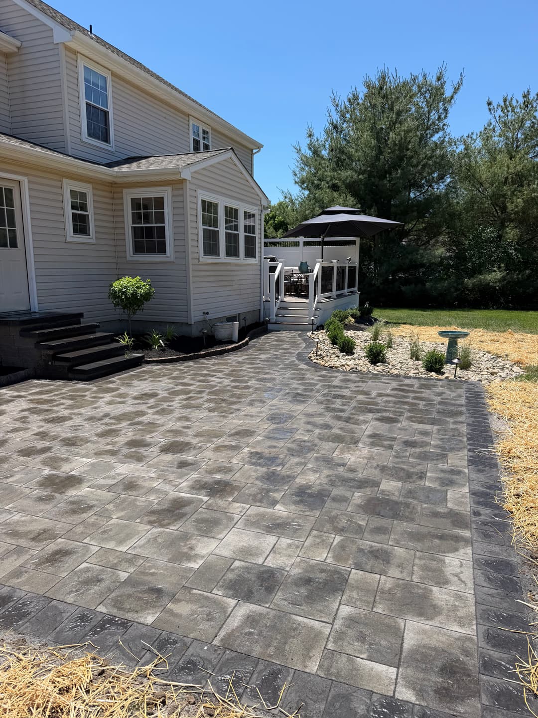 Patio with paved stones, adjacent to a house and deck, surrounded by landscaping and trees.