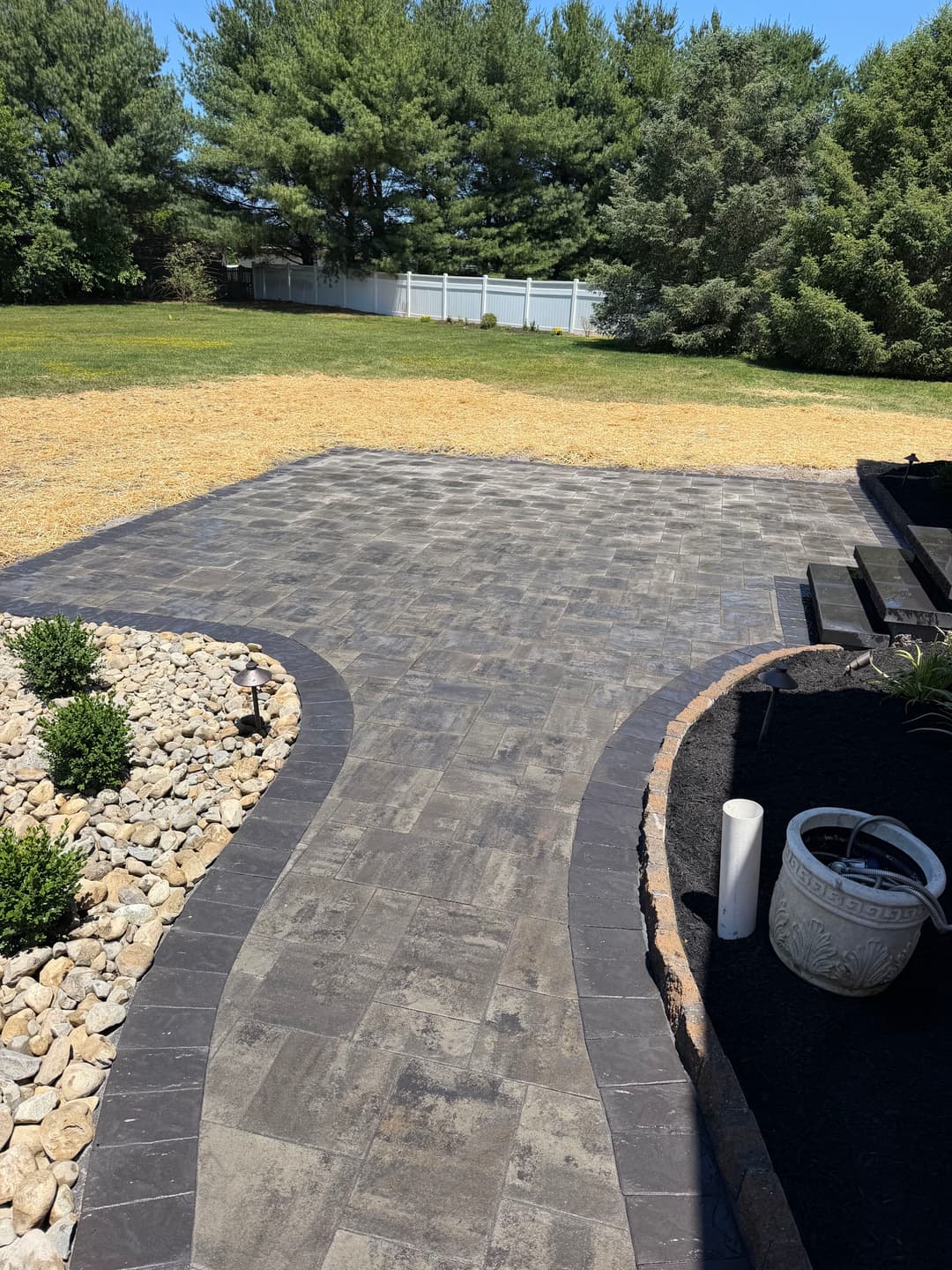 Patio with stone pavers and landscaping, bordered by grass and trees in a sunny backyard.