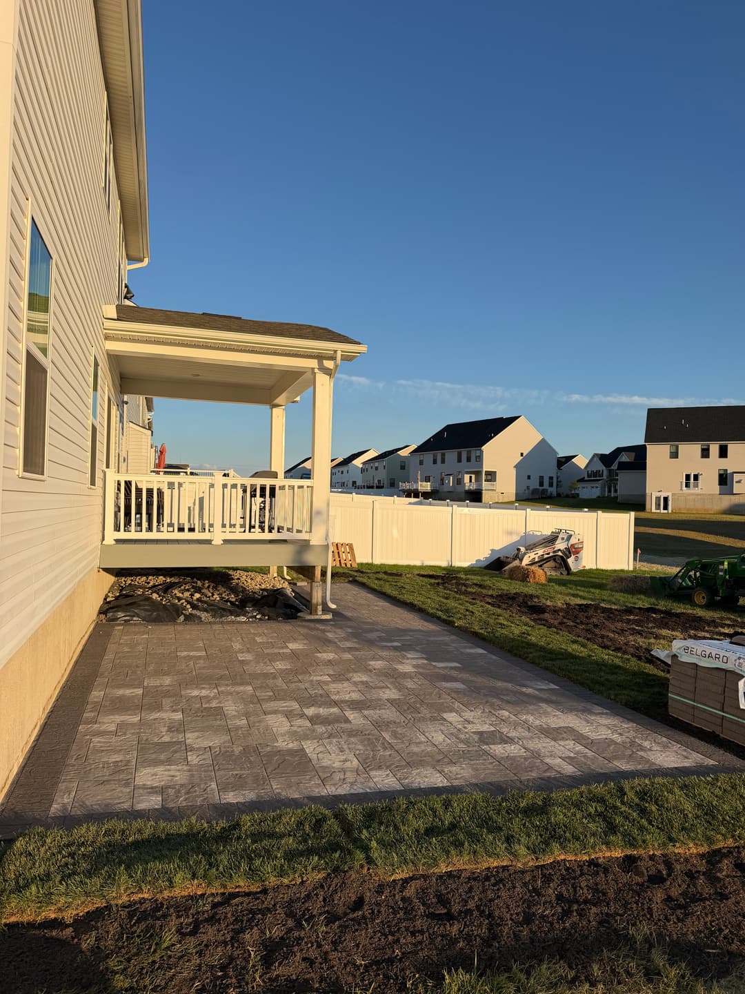 Patio area with stone pavers, grass, and a deck in a residential backyard with homes in the background.