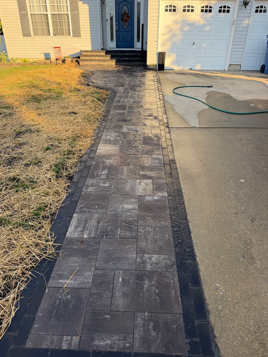 Paved walkway leading to a house with gray stone and black edging beside a driveway.