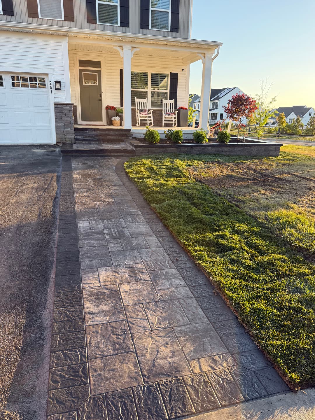 Stamped concrete walkway leading to a house entrance with landscaped yard and porch chairs.