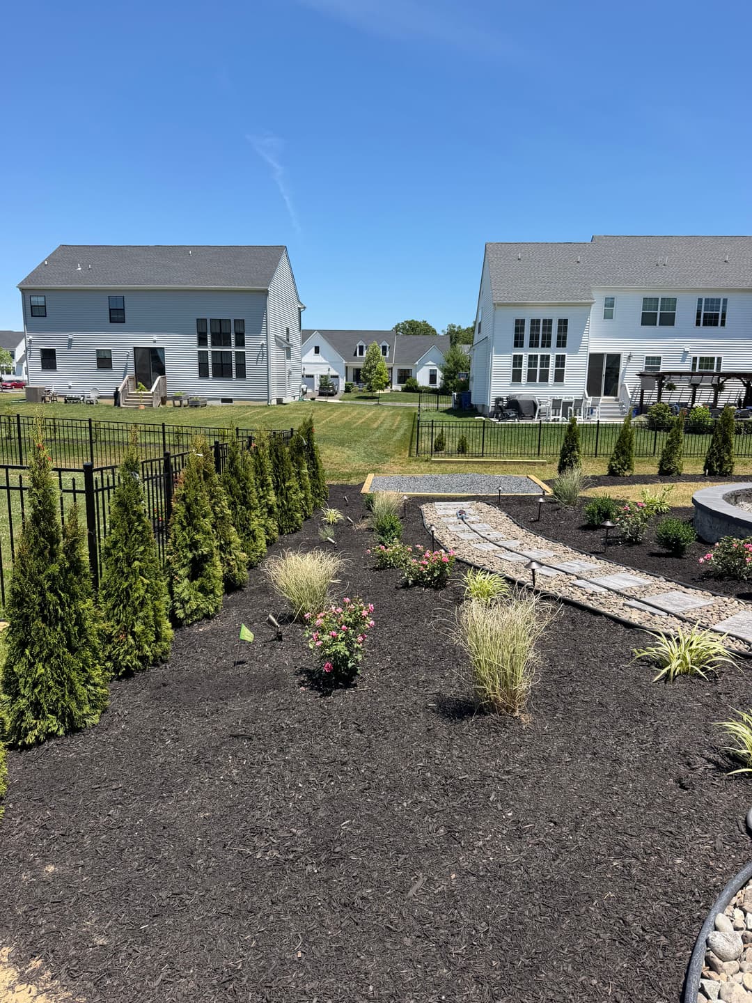 Landscaped backyard with flower beds and tall shrubs, featuring two houses in the background.