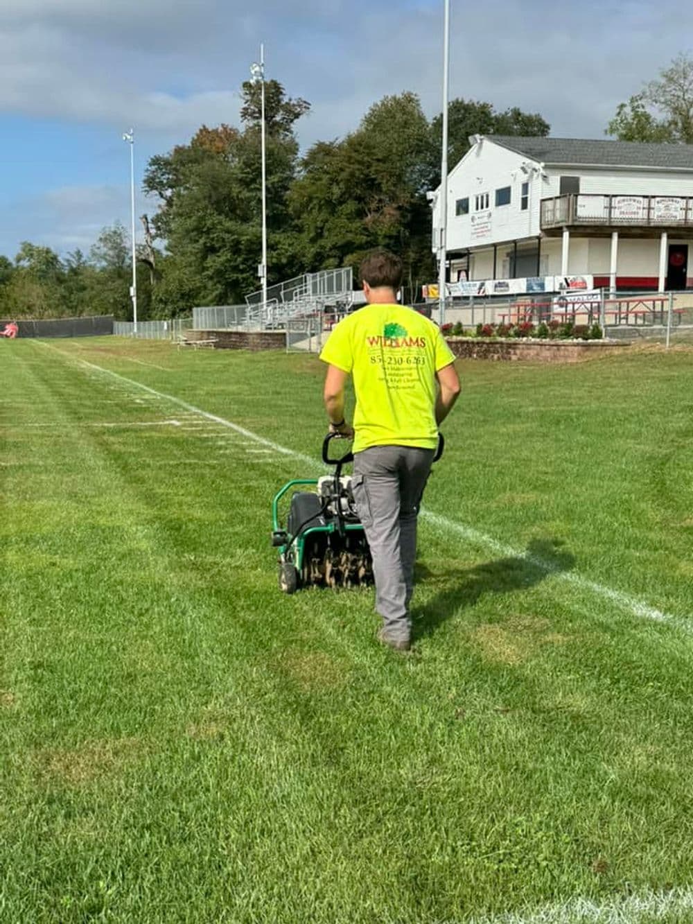 Worker aerating grass field with a power aerator, wearing a bright yellow shirt.