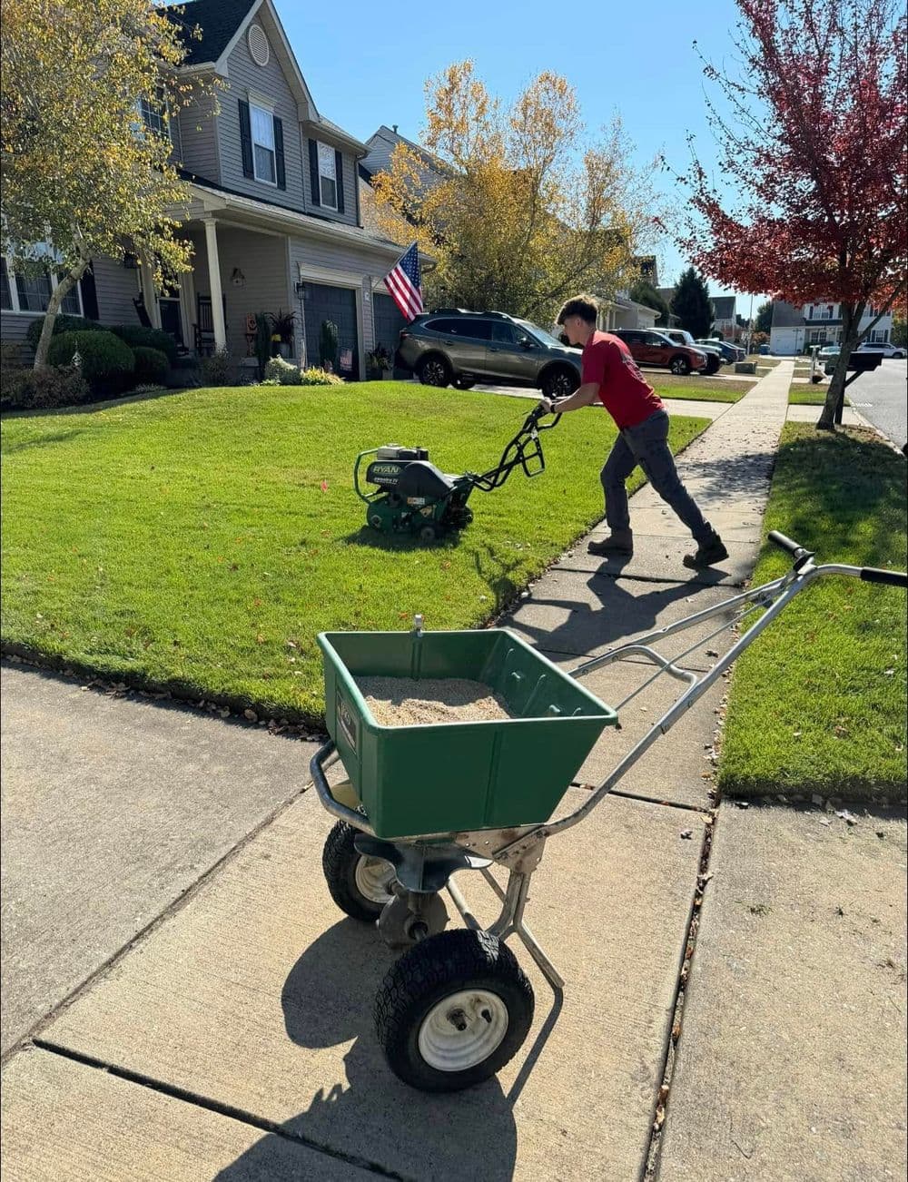 Boy mowing grass on suburban lawn with fertilizer spreader on sidewalk and American flag in background.