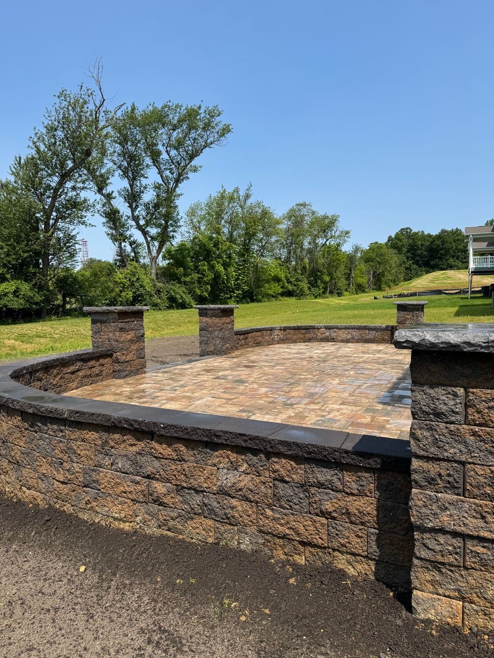 Stone patio with a retaining wall in a grassy outdoor setting under a clear blue sky.