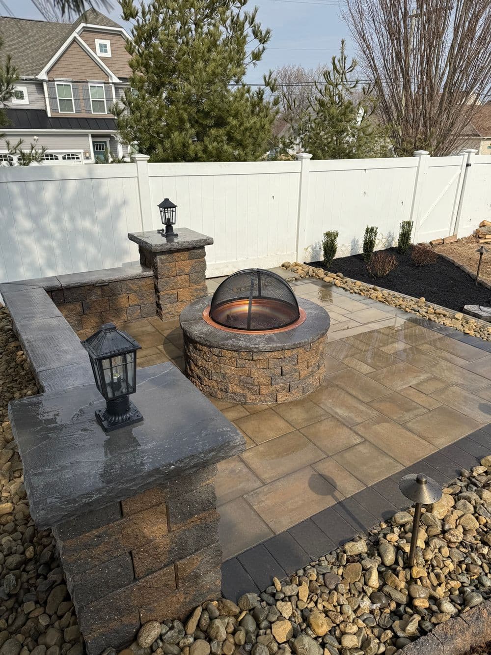 Outdoor patio with a stone fire pit, lanterns, and landscaping in a residential setting.