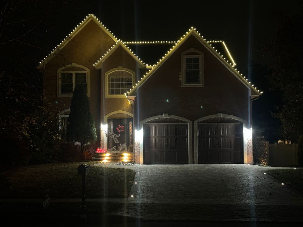 Two-story house decorated with festive white lights at night, highlighting the entrance and garage.
