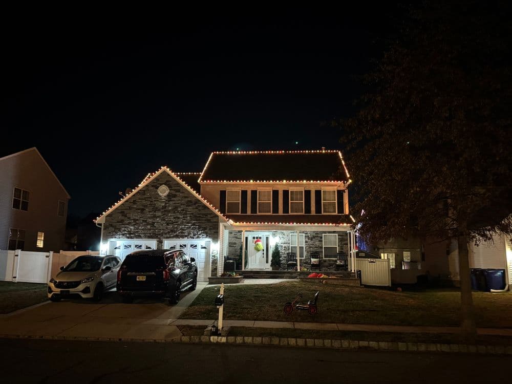Two-story house decorated with festive lights at night, surrounded by parked cars and a lawn.
