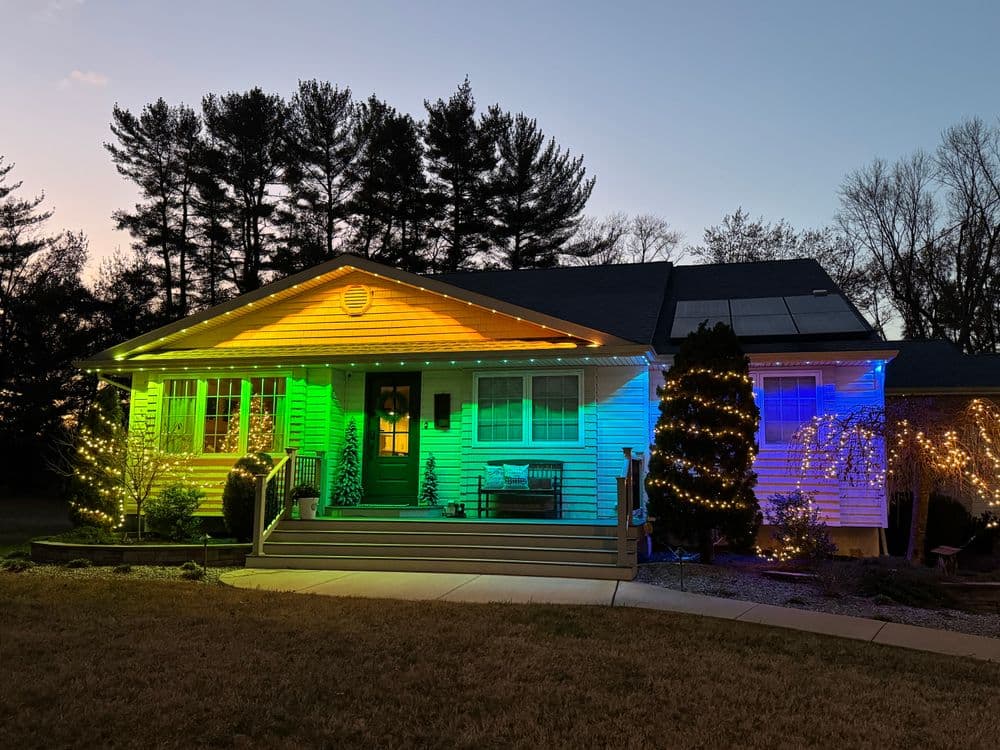 Festively lit home exterior with green and blue lights, surrounded by trees at dusk.